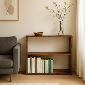 May include: A rectangular wooden console table with two open shelves. The top shelf holds a small wooden bowl, while the bottom shelf is filled with a row of books. The wood has a dark brown grain pattern and a smooth finish throughout the structure.