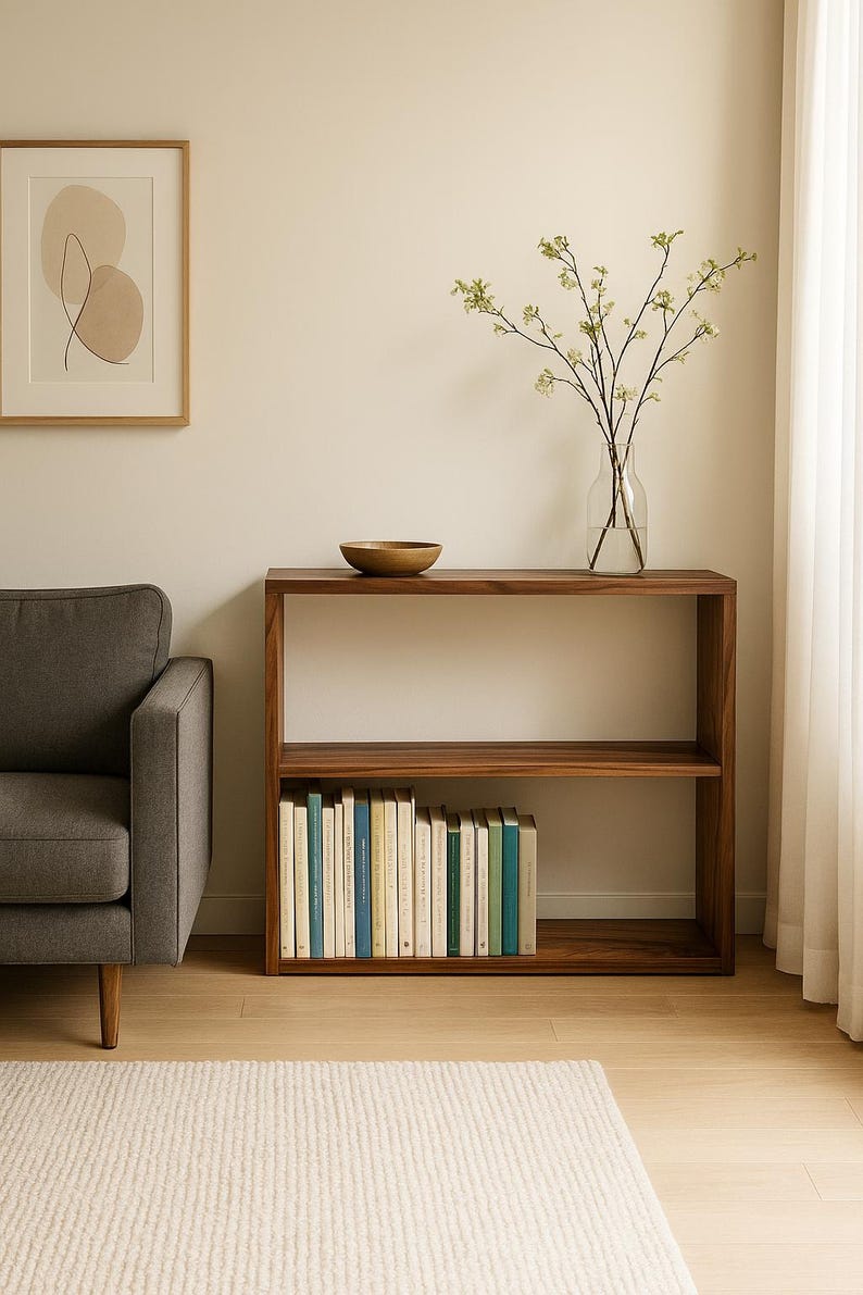 May include: A rectangular wooden console table with two open shelves. The top shelf holds a small wooden bowl, while the bottom shelf is filled with a row of books. The wood has a dark brown grain pattern and a smooth finish throughout the structure.