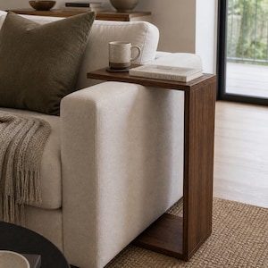 Puede incluir: Una mesa auxiliar de madera marrón oscuro con una parte superior rectangular y un marco en forma de C. Una taza y un libro están sobre la mesa. La mesa está junto a un sofá color crema con un cojín marrón y una manta beige.