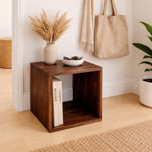 May include: A dark brown wooden cube-shaped side table with a vase of dried flowers, a white bowl, and two books. A beige tote bag and scarf hang on a wooden rack above the table. The room has light wood flooring and a woven rug.