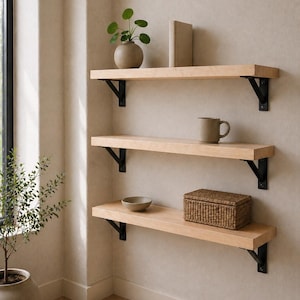 May include: Three wooden shelves mounted on a beige wall, supported by black metal brackets. The shelves hold a small potted plant, a book, a mug, a bowl, and a woven basket. The room has a neutral color palette.