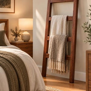 May include: A tall, dark brown wooden blanket ladder leaning against a wall. The ladder features four horizontal rungs. A cream-coloured textured throw blanket is draped over the top rung, and a tan textured throw blanket with fringe is on the second rung.