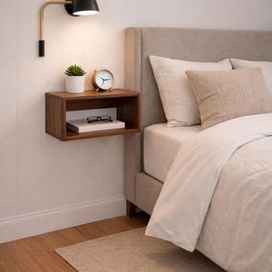 Puede incluir: Un dormitorio moderno con una mesita de noche flotante de madera que contiene una pequeña planta, un despertador dorado y unas gafas. Una lámpara de pared negra y dorada ilumina la mesita. La cama tiene un cabecero y ropa de cama de tonos neutros.