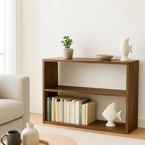 K&ouml;nnte beinhalten: Ein rechteckiges, zweist&ouml;ckiges B&uuml;cherregal aus Holz mit dunkelbrauner Oberfl&auml;che. Auf der oberen Ablage stehen eine kleine gr&uuml;ne Pflanze im Topf, eine flache beige Schale und eine wei&szlig;e Fischfigur aus Keramik. Die untere Ablage enth&auml;lt eine Reihe von B&uuml;chern.