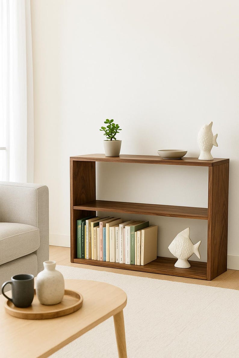 K&ouml;nnte beinhalten: Ein rechteckiges, zweist&ouml;ckiges B&uuml;cherregal aus Holz mit dunkelbrauner Oberfl&auml;che. Auf der oberen Ablage stehen eine kleine gr&uuml;ne Pflanze im Topf, eine flache beige Schale und eine wei&szlig;e Fischfigur aus Keramik. Die untere Ablage enth&auml;lt eine Reihe von B&uuml;chern.