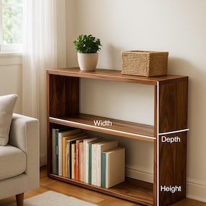 May include: A rectangular wooden bookshelf with two open shelves, featuring a dark brown wood grain finish. The unit displays white lines indicating width, depth, and height dimensions. The bottom shelf contains a row of books, while the top shelf holds a small potted green plant and a woven rectangular basket.