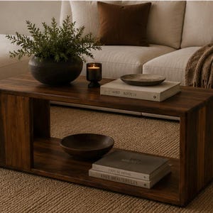 May include: A rectangular dark brown wooden coffee table with an open shelf design. The top surface holds a dark bowl with a green plant, a small lit candle in a dark glass holder, and a shallow bowl resting on a beige book. A dark bowl and two stacked books sit on the lower shelf.