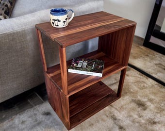 May include: A dark brown wooden side table with two shelves. A blue and white mug with a mushroom design sits on top. A book is on the middle shelf. The table is next to a gray sofa.