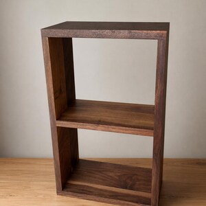 May include: A rectangular dark brown wooden shelf unit featuring a single horizontal middle shelf that divides the frame into two open storage compartments. The structure is constructed from solid wood with a visible grain pattern throughout the frame.