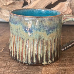 Pode incluir: Caneca de cerâmica artesanal com interior turquesa e exterior texturizado. A caneca apresenta ranhuras verticais e uma pega castanha. O exterior tem uma mistura de tons azuis, verdes e castanhos, criando um visual artesanal único.