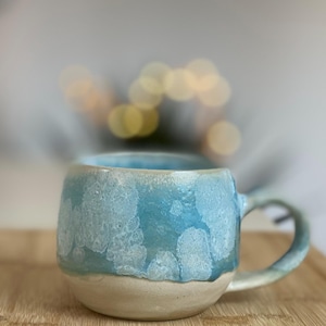 Könnte beinhalten: Handgefertigte Keramiktasse mit hellblauer Glasur und weißem, strukturiertem Muster. Die untere Hälfte der Tasse ist in einem natürlichen, unglasierten Beige gehalten. Die Tasse hat eine abgerundete Form und einen gebogenen Henkel.