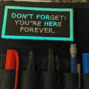 May include: Black pencil case with a teal-bordered patch that reads "DON'T FORGET! YOU'RE HERE FOREVER." The case holds a red marker, black pen, and blue pen. The case is made of a textured fabric.