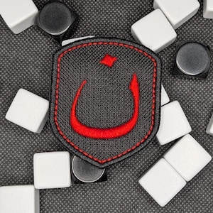 Puede incluir: Un parche negro con un diseño de media luna y punto rojo, rodeado por un borde cosido en rojo. Azulejos cuadrados blancos y imanes circulares negros dispersos sobre una superficie gris texturizada.