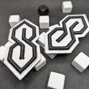 Puede incluir: Dos parches blancos bordados con un contorno negro del s&iacute;mbolo "S" estilizado. Los parches est&aacute;n rodeados de peque&ntilde;os cubos blancos y un dado negro redondeado. Los parches tienen un fondo blanco y un borde negro.
