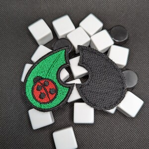May include: Two fabric patches, one green with a red ladybug and one black, are arranged on a surface of white and black tiles.