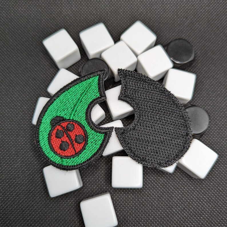 May include: Two fabric patches, one green with a red ladybug and one black, are arranged on a surface of white and black tiles.
