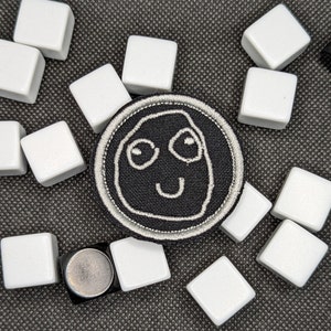 Peut inclure: Un patch brodé noir et blanc avec un visage souriant. Le patch est entouré de carreaux blancs.