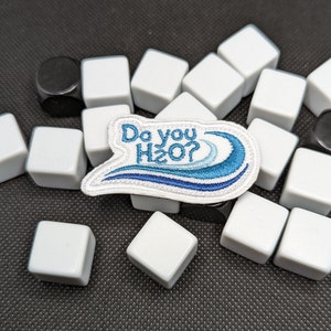 Op de afbeelding: Witte geborduurde patch met blauwe golven en de tekst "Do you H2O?" op een zwarte achtergrond met witte vierkante tegels.