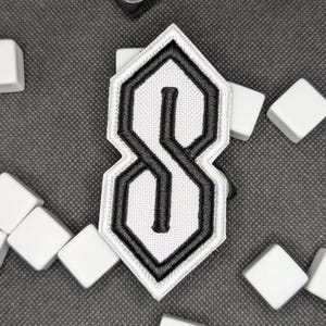 Puede incluir: Un parche bordado en blanco y negro con el ic&oacute;nico dise&ntilde;o de la "S". El parche tiene un fondo blanco con un contorno negro y una "S" negra en el centro. Azulejos cuadrados blancos rodean el parche sobre una superficie gris.