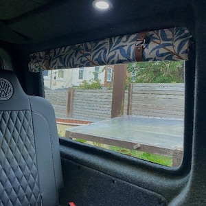 May include: A black leather car seat with a diamond pattern and a Volkswagen logo. The seat is in the back of a car with a window behind it. The window has a patterned fabric shade pulled down.