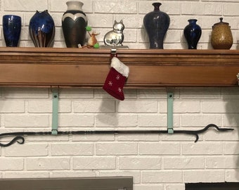 May include: A black metal fireplace mantel stocking holder with two light blue straps. The holder is attached to a white brick wall. A red stocking with white snowflakes is hanging from the holder.