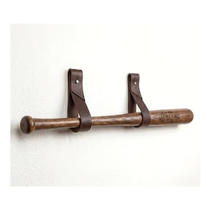 May include: A dark brown wooden baseball bat suspended horizontally by two dark brown leather straps. The bat features the text LEATHER SLINGS BAT CO. EST 1999 printed in black on the barrel. The leather straps are secured with silver rivets.