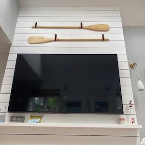 Op de afbeelding: Twee houten peddels met rode en bruine leren banden zijn gemonteerd op een witte schiplapwand boven een zwarte flatscreen-televisie. De televisie is gemonteerd op een witte schouw.