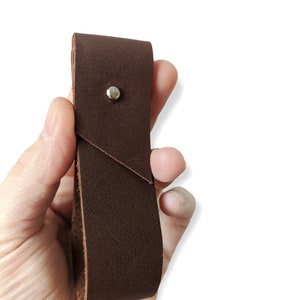 Op de afbeelding: Een bruine leren lus met een zilveren klinknagel. De lus is dubbelgevouwen en heeft een glad, gestructureerd oppervlak.