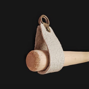 May include: A white leather strap with a brass ring and a wooden dowel attached. The strap has a textured surface and is designed to hang an object.