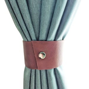 Op de afbeelding: Een bruine leren gordijn terugslag met een zilveren drukknop, gewikkeld rond een gedeelte van geplooide, lichtblauwe gordijnen. De terugslag heeft een eenvoudig, maar elegant ontwerp.