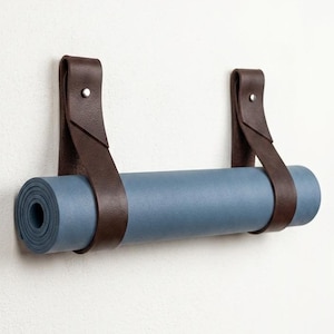 May include: A rolled blue yoga mat suspended horizontally by two dark brown leather straps. Each strap is secured to the wall with a single silver-toned metal rivet, creating a loop that holds the mat firmly in place against the flat surface.