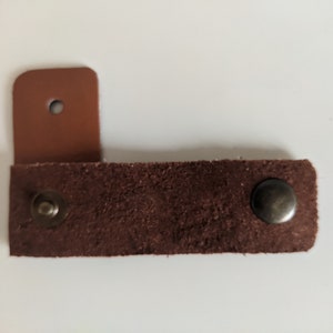 May include: A brown leather strap with two metal snaps. The strap is attached to a smaller piece of brown leather with a hole in the center.