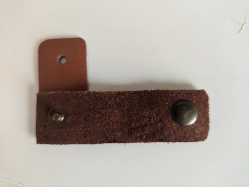 May include: A brown leather strap with two metal snaps. The strap is attached to a smaller piece of brown leather with a hole in the center.