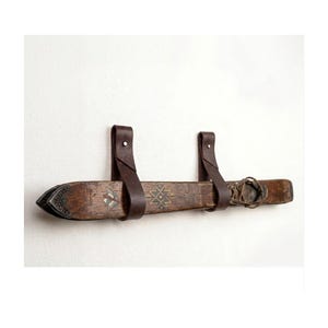 May include: A pair of vintage wooden skis mounted horizontally on a wall using two dark brown leather straps. The skis feature a weathered brown finish with decorative geometric patterns and a small bird graphic, secured by metal rivets on the straps.