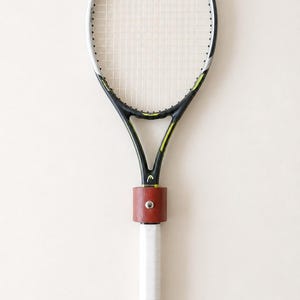 May include: A black and yellow HEAD tennis racket with white strings and a white grip. A brown leather band is secured around the throat of the racket with a silver metal stud. The word HEAD is printed in white at the top and bottom of the frame.