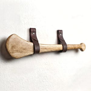 Könnte beinhalten: Ein hölzerner Hurling-Schläger, der horizontal mit zwei dunkelbraunen Lederriemen an einer Wand befestigt ist. Der Schläger hat eine natürliche helle Holzmaserung, einen breiten, flachen Kopf links und einen runden Knauf am Griffende.