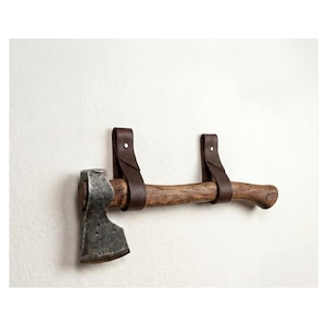 May include: A hand axe with a weathered metal head and a textured wooden handle, suspended horizontally by two dark brown leather straps that are secured with silver-toned metal rivets, creating a wall-mounted display arrangement.
