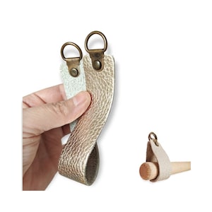 Puede incluir: Dos correas de cuero con anillos y remaches de metal color latón. Una correa es dorada, la otra verde claro. Las correas se muestran en una mano y también unidas a un pasador de madera. El cuero tiene una apariencia texturizada.