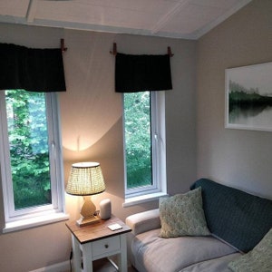 Op de afbeelding: Een woonkamer met een bank, twee ramen met zwarte gordijnen, een bijzettafel met een lamp en een ingelijst schilderij aan de muur.