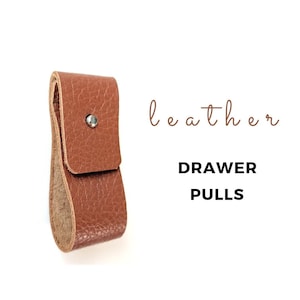 May include: A brown leather drawer pull with a silver rivet. The text "leather" and "DRAWER PULLS" is printed below the pull.