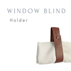 May include: A brown leather strap with a metal button wraps around a white fabric window blind. The strap is designed to hold the blind in place when it is rolled up. The text "WINDOW BLIND Holder" is written above the blind.