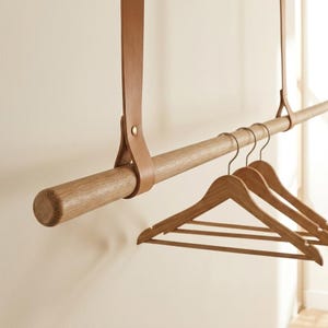 May include: A horizontal wooden rod suspended by two tan leather straps with brass-coloured metal rivets. Three wooden clothes hangers are hooked onto the rod. The rod has a visible wood grain texture and rounded ends, providing a hanging storage solution.