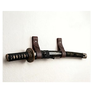 Könnte beinhalten: Ein Katana-Schwert mit schwarz umwickeltem Griff und dunkel gemusterter Scheide, das horizontal von zwei braunen Lederriemen gehalten wird. Das Schwert verfügt über ein rundes Metall-Parierelement und eine silberfarbene Endkappe.