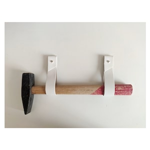 May include: A white leather hammer holder with two straps attached to a wall. A black headed hammer with a wooden handle is hanging from the straps.