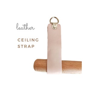 May include: A light pink leather ceiling strap with a silver metal ring and a silver metal button. The strap is attached to a wooden dowel.