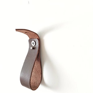 Op de afbeelding: Een bruine leren haak gemonteerd op een witte muur. De haak is lusvormig en heeft een metalen schroef.