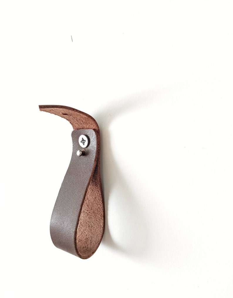 Op de afbeelding: Een bruine leren haak gemonteerd op een witte muur. De haak is lusvormig en heeft een metalen schroef.