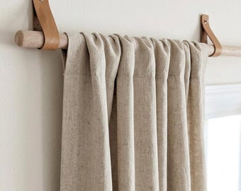 Könnte beinhalten: Ein beigefarbener Leinenvorhang hängt an einer hellen Holzstange. Die Stange wird von zwei hellbraunen Lederriemen gehalten, die mit silberfarbenen Metallbeschlägen an der Wand befestigt sind, was eine einfache Fensterdekoration ergibt.