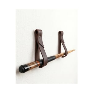 Könnte beinhalten: Zwei dunkelbraune Lederriemen sind an einer Wand befestigt und bilden Schlaufen, die ein hölzernes Billard-Queue halten. Die Lederriemen sind mit silberfarbenen Metallnieten gesichert und bieten ein stabiles Haltesystem für den horizontalen Stock.
