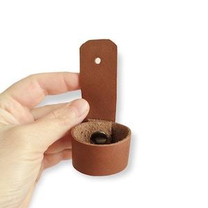 May include: A brown leather strap with a hole and a snap closure. The strap is folded in half to show the inside of the strap.